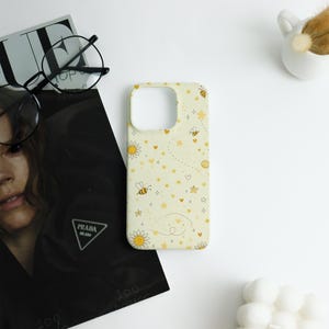 May include: A pale yellow phone case featuring a pattern of bees, daisies, stars, and hearts. The case has a camera and button cutouts. The design uses yellow and orange hues.