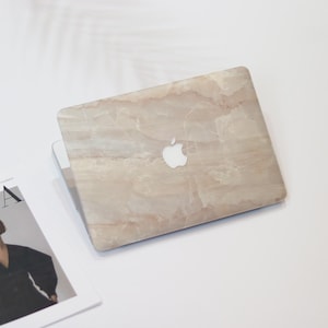 Könnte beinhalten: Ein Laptop mit marmorähnlichem Design auf der Abdeckung, mit einem weißen Apple-Logo. Die Abdeckung hat ein beige-cremefarbenes Farbschema mit subtilen Maserungen. Eine Zeitschrift ist in der linken unteren Ecke sichtbar.