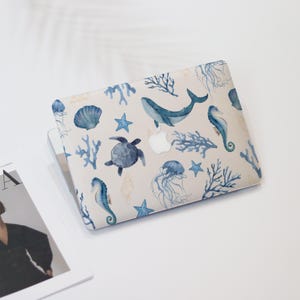 May include: A laptop case with a marine-themed watercolour design. The case features illustrations of a whale, seahorse, jellyfish, and sea turtle. The background is light beige with blue and white coral, starfish, and seashells.