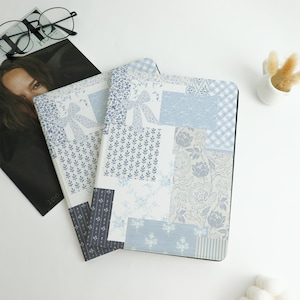May include: Two notebooks with a patchwork design in shades of blue and white. The covers feature floral, bow, and gingham patterns. A magazine and eyeglasses are in the background.