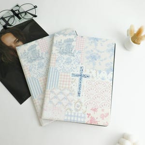 May include: Two notebooks with a patchwork design in pastel colors, featuring floral and toile patterns, and a blue cross. The notebooks are on a white surface, with a magazine and glasses in the background.