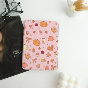 Könnte beinhalten: Eine rosa Tablet-Hülle mit einem sich wiederholenden Muster aus Kürbissen, Pullovern, Kaffeetassen und anderen herbstlichen Illustrationen. Die Hülle hat abgerundete Ecken und eine glatte Oberfläche.