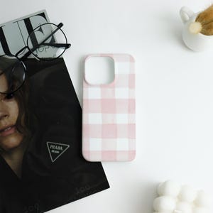 May include: A pink and white gingham pattern phone case. The case is made of a hard plastic material and has a rectangular shape with rounded corners. The case is next to a black magazine, a pair of glasses, and a small white ceramic pot.