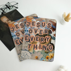 Peut inclure: Deux cahiers avec le texte "JESUS OVER EVERYTHING" en lettres orange et noires. Les couvertures présentent un collage d'éléments floraux et de journaux. Un magazine et des lunettes sont visibles en arrière-plan.