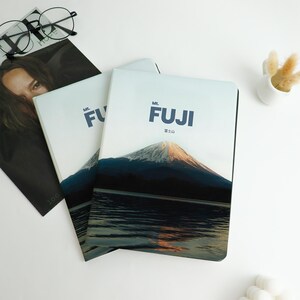 May include: Three notebooks with the image of Mount Fuji on the cover. The notebooks have a light blue background with the mountain and lake in the foreground. The text "Mt. FUJI" is printed in dark blue. A magazine and glasses are in the background.