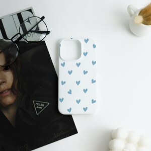 May include: White phone case with a pattern of small, light blue hearts. The case is shown on a white surface next to a magazine, eyeglasses, and a small white ceramic container.