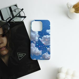 May include: A blue phone case featuring a sky design with white clouds and colorful stars. The case has a glossy finish and is designed to fit a smartphone. The case is shown on a white surface with other objects.