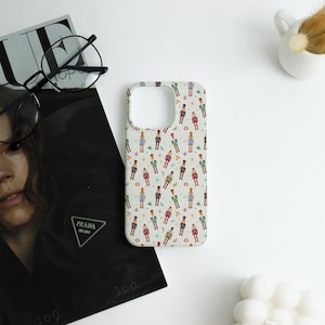 May include: A phone case with a repeating pattern of colorful toy soldiers on a light beige background. The design includes red, green, blue, and black soldiers, along with stars and circles. The case is shown on a white surface.