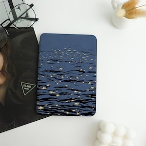 May include: A rectangular, navy blue case featuring a stylized illustration of a starry night sky. The design includes gold stars and dark blue water-like lines. The case is likely for a phone or tablet.