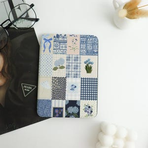 May include: A square item with a patchwork design. The design features various floral and geometric patterns in shades of blue, white, and beige. The item is likely a decorative piece or a functional item like a coaster or a phone case.