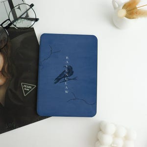 May include: A navy blue notebook featuring a black raven illustration and the word "Ravenclaw" printed vertically in white. The notebook has a minimalist design with decorative lines.