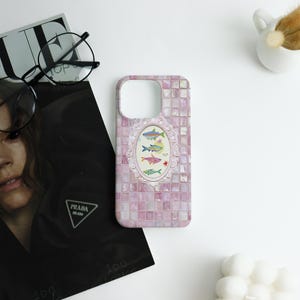 May include: A pink phone case with a mosaic tile design. The case features an oval frame with illustrations of colorful fish. The background includes a magazine, glasses, and a small white vase.