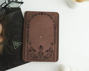 May include: A brown journal with an ornate black design. The cover features an arched frame with floral and geometric patterns. A small gold-colored star is in the center of the cover. The journal is rectangular and appears to be closed.