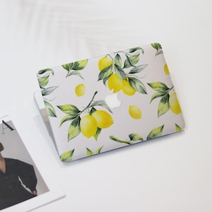 Könnte beinhalten: Ein Laptop mit weißer Hülle, verziert mit einem Aquarellmuster aus gelben Zitronen und grünen Blättern. Das Muster wiederholt sich auf der gesamten Hülle, mit dem Apple-Logo in der Mitte.
