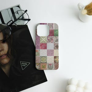 May include: A phone case featuring a patchwork design of various square tiles in shades of pink, green, and cream. The tiles display floral and geometric patterns. The case is on a white surface with a magazine, glasses, and a small decorative item.