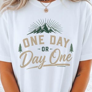 Op de afbeelding: Wit T-shirt met een berg- en boomontwerp. De tekst "ONE DAY -or- Day One" is gedrukt in groen en beige. Het T-shirt heeft een casual stijl.