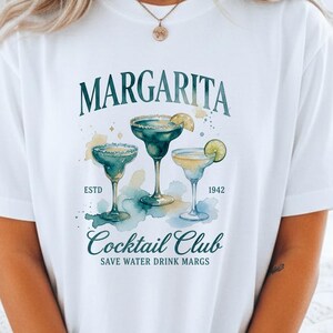 May include: White t-shirt featuring a watercolor design of three margarita cocktails. The shirt has the text "Margarita Cocktail Club" with the tagline "Save Water Drink Margs." The design includes the year "1942" and the word "ESTD".