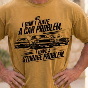 Puede incluir: Camiseta de color amarillo mostaza con el texto "NO, I DON'T HAVE A CAR PROBLEM. I HAVE A STORAGE PROBLEM." Tres siluetas de coches negros están impresas debajo. La camiseta la lleva una persona.