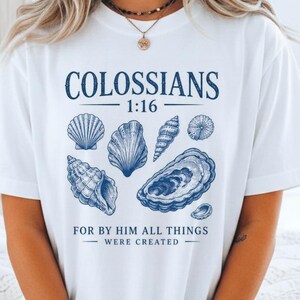 May include: White t-shirt featuring a blue graphic of various seashells and the text "COLOSSIANS 1:16 FOR BY HIM ALL THINGS WERE CREATED". The design is centered on the front of the shirt.
