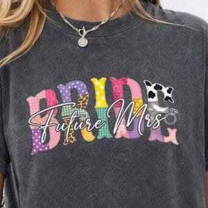 May include: Dark gray t-shirt with the words "BRIDE Future Mrs" in a colorful patchwork design. The word "BRIDE" is in large, bold letters, and "Future Mrs" is in a script font. The shirt is a casual style.