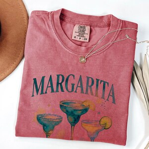 May include: A dusty rose-colored t-shirt with the word "MARGARITA" in teal lettering. Below the text are three margarita glasses in teal and orange. A silver necklace with a pendant is draped across the shirt.
