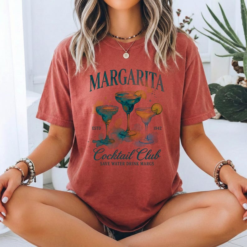 May include: A brick-red t-shirt featuring the text "Margarita Cocktail Club" with illustrations of three margarita cocktails. The shirt includes the text "Save Water Drink Margs" and the years "ESTD 1942".