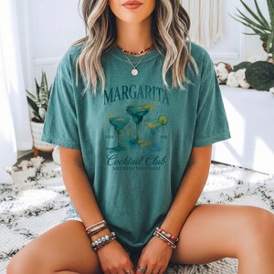 May include: Teal-colored t-shirt with the word "Margarita" above a graphic of three margarita cocktails. The shirt also features the text "Cocktail Club" and "Save Water Drink Margs".