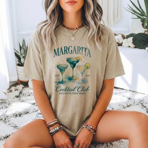 May include: A tan t-shirt featuring the word "Margarita" above a watercolor illustration of three margarita cocktails. The shirt also includes the text "Cocktail Club" and "Save Water Drink Margs."