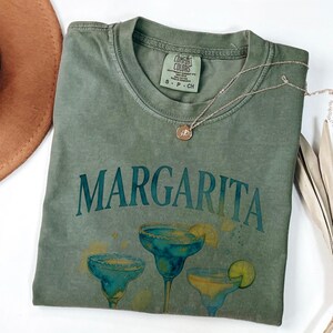 May include: Green t-shirt with the word "Margarita" in teal lettering and illustrations of margarita cocktails. The shirt has a crew neck and short sleeves. A silver necklace with a pendant is draped across the shirt.