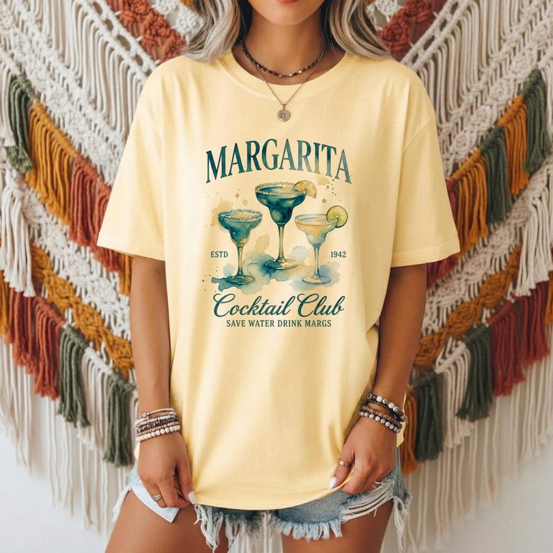 May include: Yellow t-shirt with a Margarita Cocktail Club design. The design features three margarita glasses with lime wedges, the word "Margarita" above, and the text "Save Water Drink Margs" below. The shirt is a light yellow color.