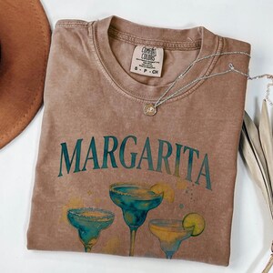May include: Brown t-shirt with the word "Margarita" in teal lettering and a watercolor illustration of three margarita cocktails. A silver necklace and a brown hat are also visible.