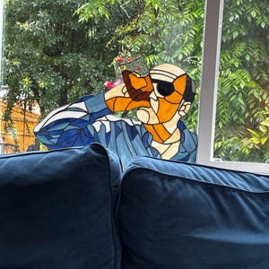 May include: A stained glass panel depicting a person wearing dark sunglasses and a blue shirt, holding a glass to their mouth. The piece features orange, blue, white, and black glass segments joined by dark lead lines, creating a stylised portrait.
