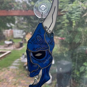May include: A stained glass suncatcher shaped like a blue oni mask with a single white horn. The mask features black line details, a dark eye socket, and sharp white teeth. It is suspended by a clear suction cup attached to a metal ring at the top.