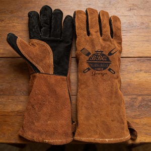 May include: A pair of brown leather BBQ gloves with black accents. The right glove features a black graphic with the words "BBQ Master" and the name "George". The gloves are designed for grilling and outdoor cooking.