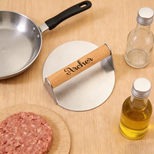May include: A stainless steel burger press with a wooden handle, engraved with the name "Archer", sits on a wooden surface. A small frying pan, a raw burger patty, and two small glass bottles are also present.