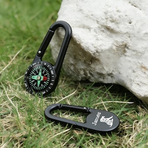 May include: Black compass with a carabiner clip next to a matching black clip with the name "James" and a dog graphic. The compass has a black bezel with white and green markings. The items are on green grass.
