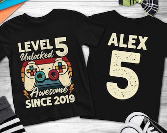 May include: Two black t-shirts. One features a retro video game controller graphic with the text "LEVEL 5 Unlocked Awesome SINCE 2019". The other shirt has the name "ALEX" and the number "5".