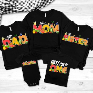 May include: Black t-shirts and a onesie with colorful text and fruit graphics. The shirts say "DAD", "MOM", and "SISTER". The onesie says "BROTHER", and a shirt says "HEY! I'M ONE".
