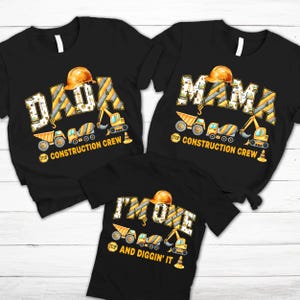 May include: Black t-shirts with construction-themed graphics. One shirt says "DADA" with a hard hat and construction vehicles. Another says "MAMA". The third shirt says "I'M ONE" and "AND DIGGIN' IT".