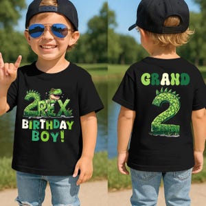 May include: Black t-shirt with a green dinosaur graphic and the text "2 Rex Birthday Boy!" on the front. The back of the shirt has the word "Grand" and the number "2" in green. The shirt is worn by a child.