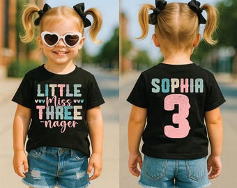 May include: A black t-shirt with the text "LITTLE Miss THREE-nager" in white, pink, and teal. The back of the shirt has the name "SOPHIA" and the number "3" in pink. The child is wearing denim shorts and heart-shaped sunglasses.