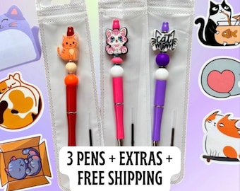 May include: Three pens with cat-themed toppers in clear plastic packaging. One pen is red with an orange cat, one is pink with a pink cat, and one is purple with "cat mom" text. Cat stickers surround the pens.