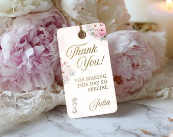 以下が含まれることがあります： 金色の筆記体で「Thank You！」と書かれた長方形のギフトタグ。 「For making this day so special」というテキストと「Julia」という名前も含まれています。 タグはピンクの花柄のアクセントと金色の紐で飾られています。