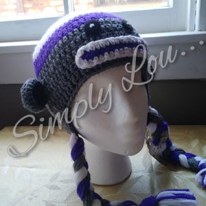May include: A handmade crochet hat in the shape of a monkey, featuring gray, purple, and white yarn. The hat has a round head, a white and purple mouth, and braided ear flaps. The text "Simply Lou" is written on the image.
