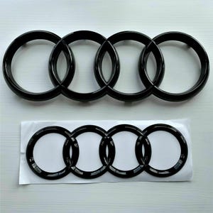 May include: Two black Audi emblem logos. One is a standalone emblem, and the other is an emblem on a white backing. The emblems feature four interlocking rings, a signature design element of the Audi brand.