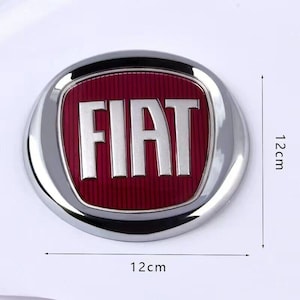 May include: A silver and red car emblem with the word "FIAT" in white letters. The emblem is 12 cm in diameter.
