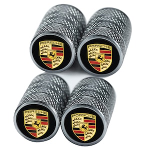 May include: A collection of silver-coloured tyre valve stem caps with a diamond-patterned texture. Each cap displays a black circular emblem with the Porsche logo in gold, red, and black. These caps are designed for automotive use.