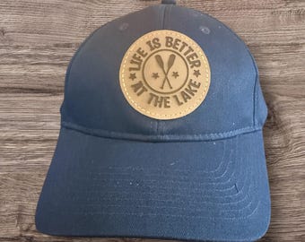 May include: A navy blue baseball cap with a curved brim. A circular patch on the front reads "LIFE IS BETTER AT THE LAKE" with crossed oars. The patch is tan with a star on each side.