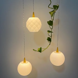 Könnte beinhalten: Drei Pendelleuchten mit weißen Schirmen und gelben Akzenten hängen von der Decke. Die Schirme haben unterschiedliche Texturen: einer mit einem Rautenmuster und zwei mit vertikalen Linien. Eine grüne Rebe mit Blättern ist ebenfalls aufgehängt.