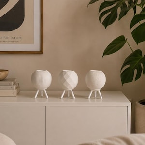 Könnte beinhalten: Drei weiße, dekorative, kugelförmige Vasen mit strukturierten Oberflächen und geometrischen Designs, jede von drei kleinen weißen Beinen getragen. Die Vasen stehen auf einem weißen Schrank, im Hintergrund ein gerahmter Kunstdruck und eine Topfpflanze.